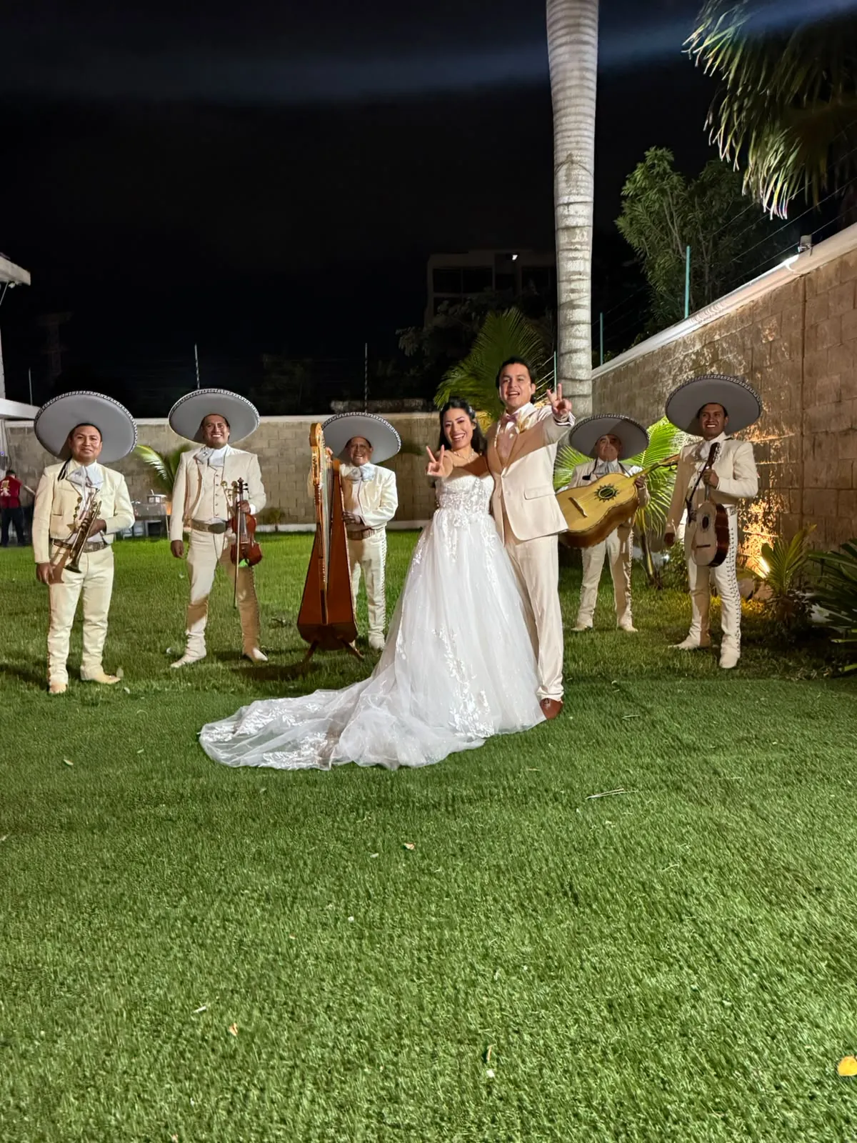 Novios celebrando con seis músicos de mariachi y un arpa sobre el césped de un jardín de noche