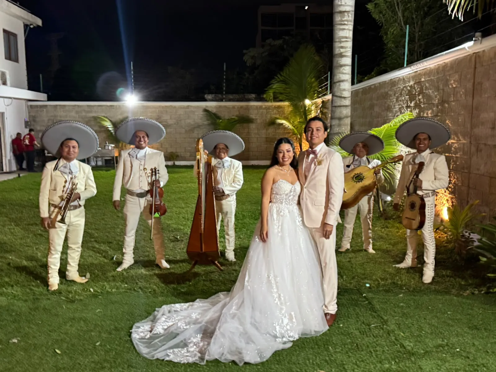Novios posando con un mariachi de seis músicos y arpa durante una boda en jardín por la noche