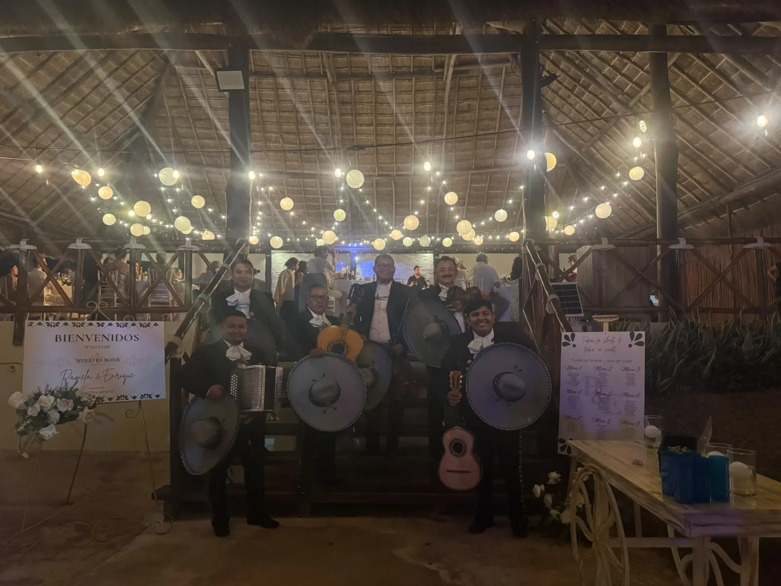 Mariachi con acordeón y sombreros posando en una boda bajo una palapa con luces cálidas