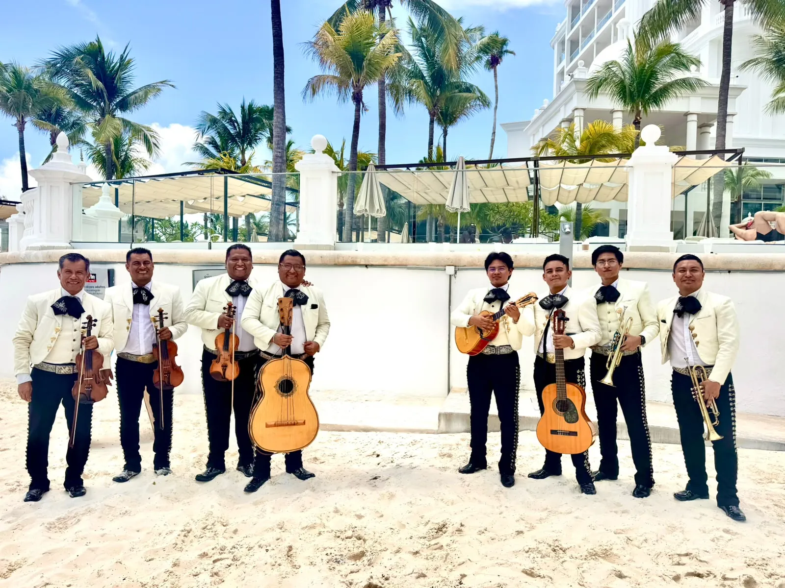 Ocho músicos de mariachi con saco claro posando en la arena frente a un hotel en la playa