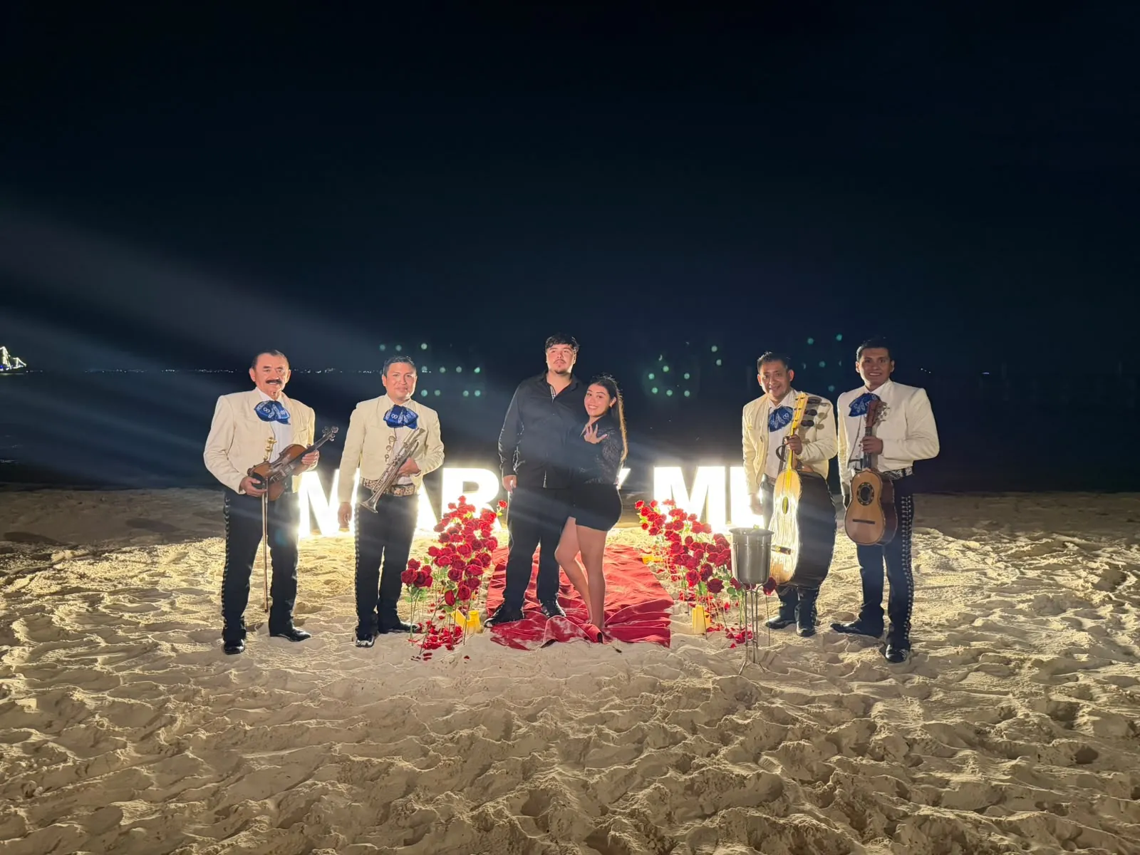 Pareja celebrando una propuesta de matrimonio de noche en la playa, con mariachi, letras iluminadas y camino de rosas rojas